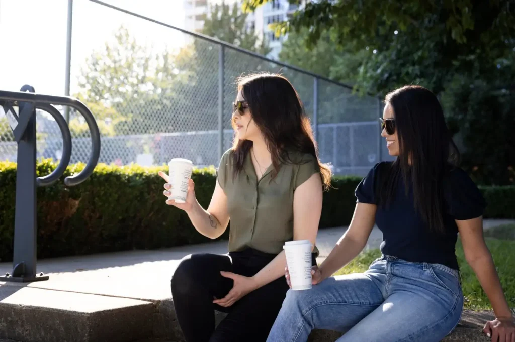 Two women sit on a step outdoors, holding coffee cups, wearing sunglasses, and talking mid-conversation, with greenery behind them.