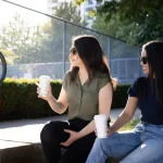 Two women sit on a step outdoors, holding coffee cups, wearing sunglasses, and talking mid-conversation, with greenery behind them.