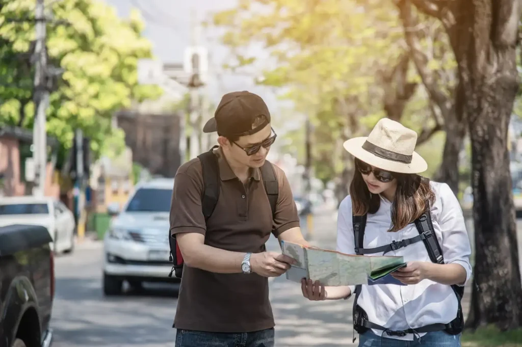 Two travelers with backpacks looking at a map on a sunny street lined with trees and parked cars.