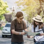 Two travelers with backpacks looking at a map on a sunny street lined with trees and parked cars.