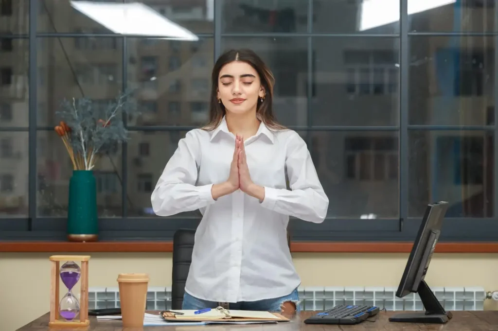 Woman in a white shirt practices meditation with hands in prayer at her desk in an office with large windows.