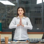 Woman in a white shirt practices meditation with hands in prayer at her desk in an office with large windows.