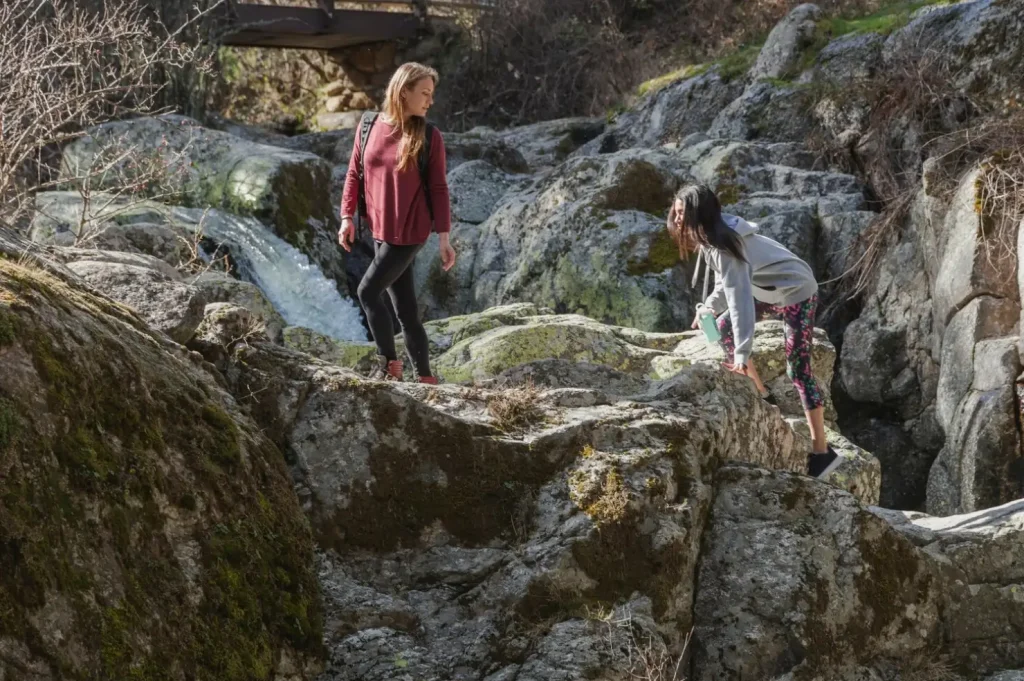 Two women hiking and climbing over large moss-covered rocks near a waterfall and a wooden bridge in a forested area.