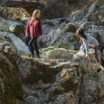 Two women hiking and climbing over large moss-covered rocks near a waterfall and a wooden bridge in a forested area.