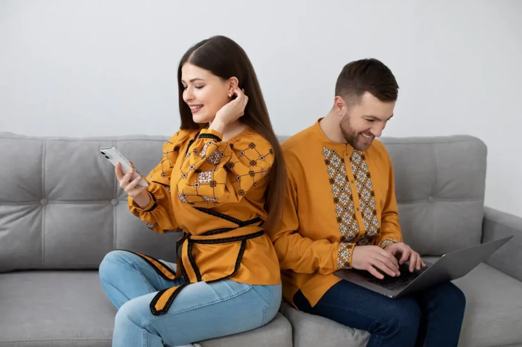 Two people on a gray couch, wearing matching yellow embroidered tops; one using a smartphone, the other a laptop.