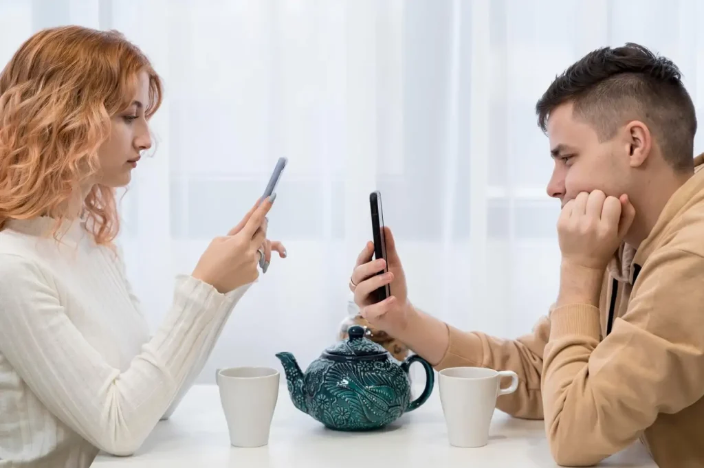 Two people sit at a table with a teal teapot and two white mugs, both looking at their smartphones.