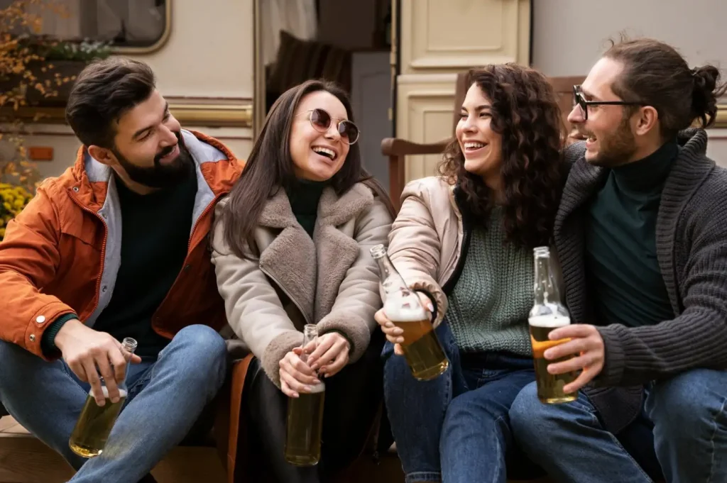 Four friends sit outdoors, laughing and holding beers, dressed warmly in jackets and sweaters, enjoying a casual gathering.