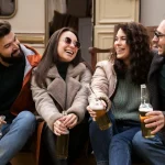 Four friends sit outdoors, laughing and holding beers, dressed warmly in jackets and sweaters, enjoying a casual gathering.