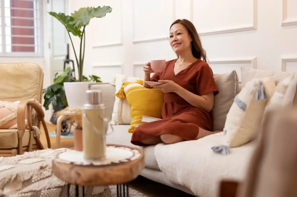 Woman in a red dress sits on a beige sofa, holding a pink cup and saucer, with pillows and a thermos in a cozy living room.