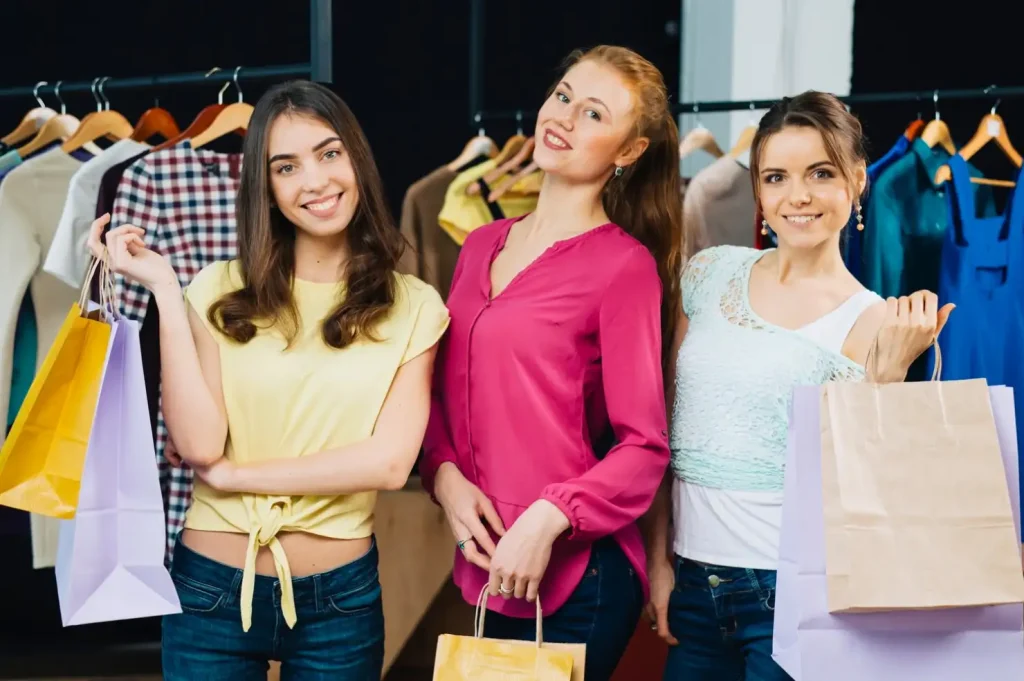 Three women with shopping bags smiling in a clothing store. Various outfits on hangers are displayed in the background.