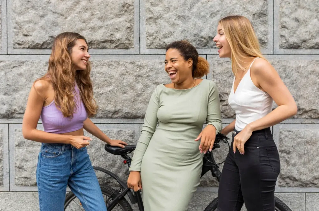Three women laughing and chatting while leaning on a bike against a stone wall.