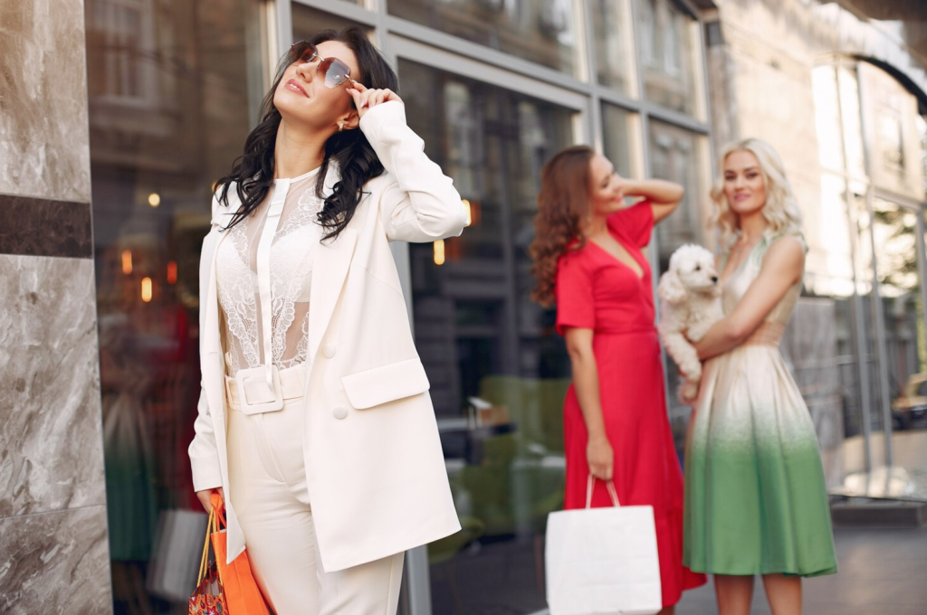 Three women, dressed stylishly, socializing outdoors. One adjusts sunglasses, two others chat, one holding a small dog. Shopping bags visible.