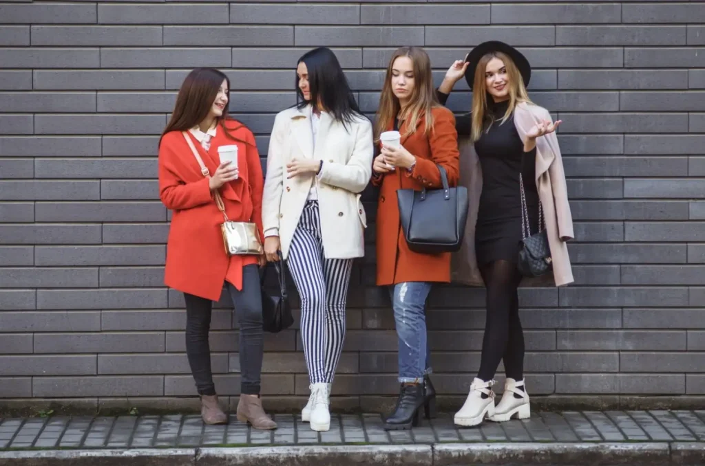 Four fashionable women in stylish outfits, standing against a grey brick wall, holding coffee cups and chatting.