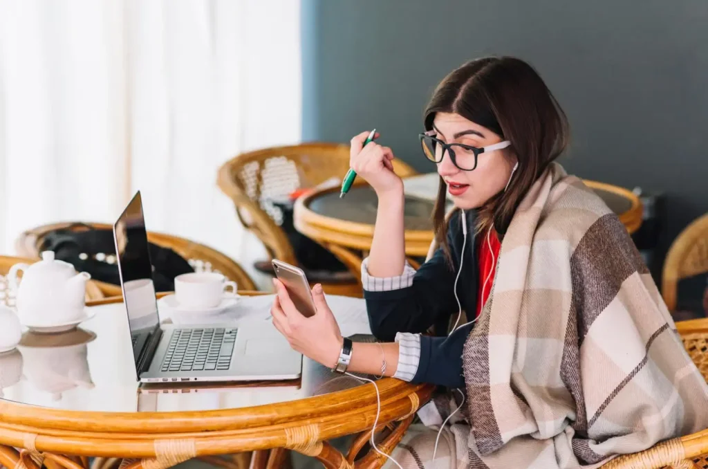 Woman in glasses, holding a pen, and wrapped in a blanket, speaks on her phone while working on a laptop at a café table.